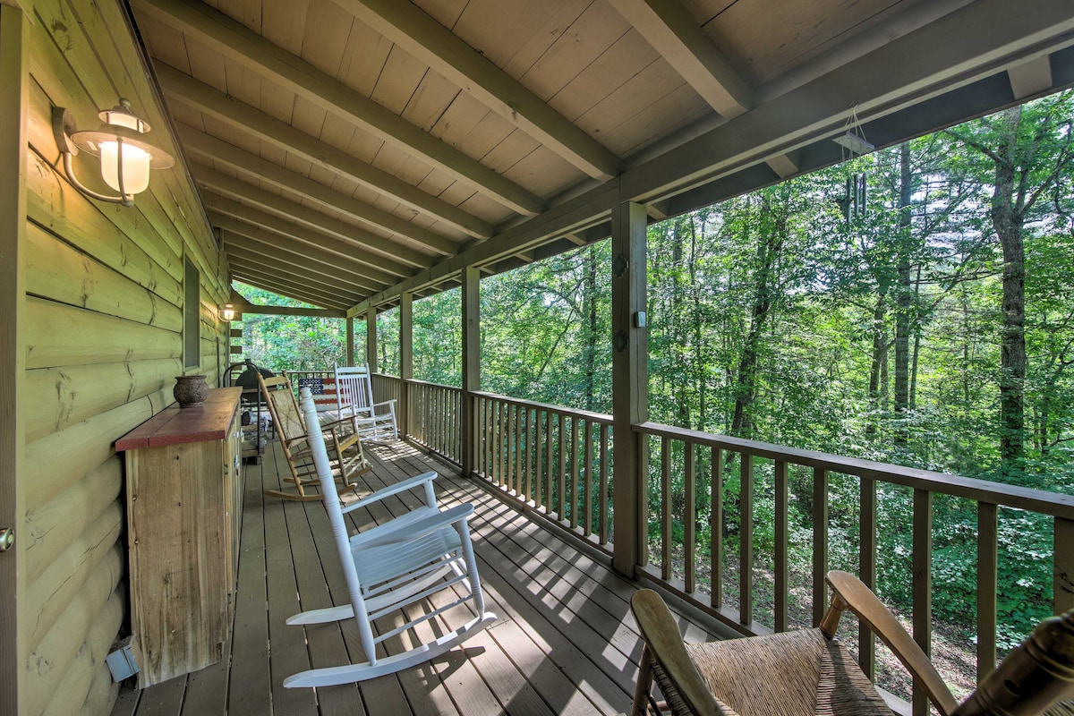A covered wraparound deck is shown, featuring multiple rocking chairs and wooden railings. The surrounding forest provides a natural backdrop, while soft lighting fixtures add an inviting touch to the outdoor space.