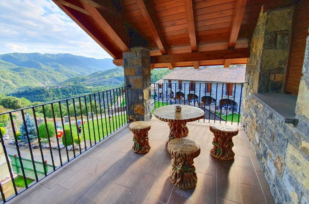 A private terrace is furnished with a round table and four unique wooden stools, designed to blend with the natural surroundings. The wooden beams and stone walls create a rustic ambiance, while expansive views of the mountains and landscaped gardens are visible in the background.