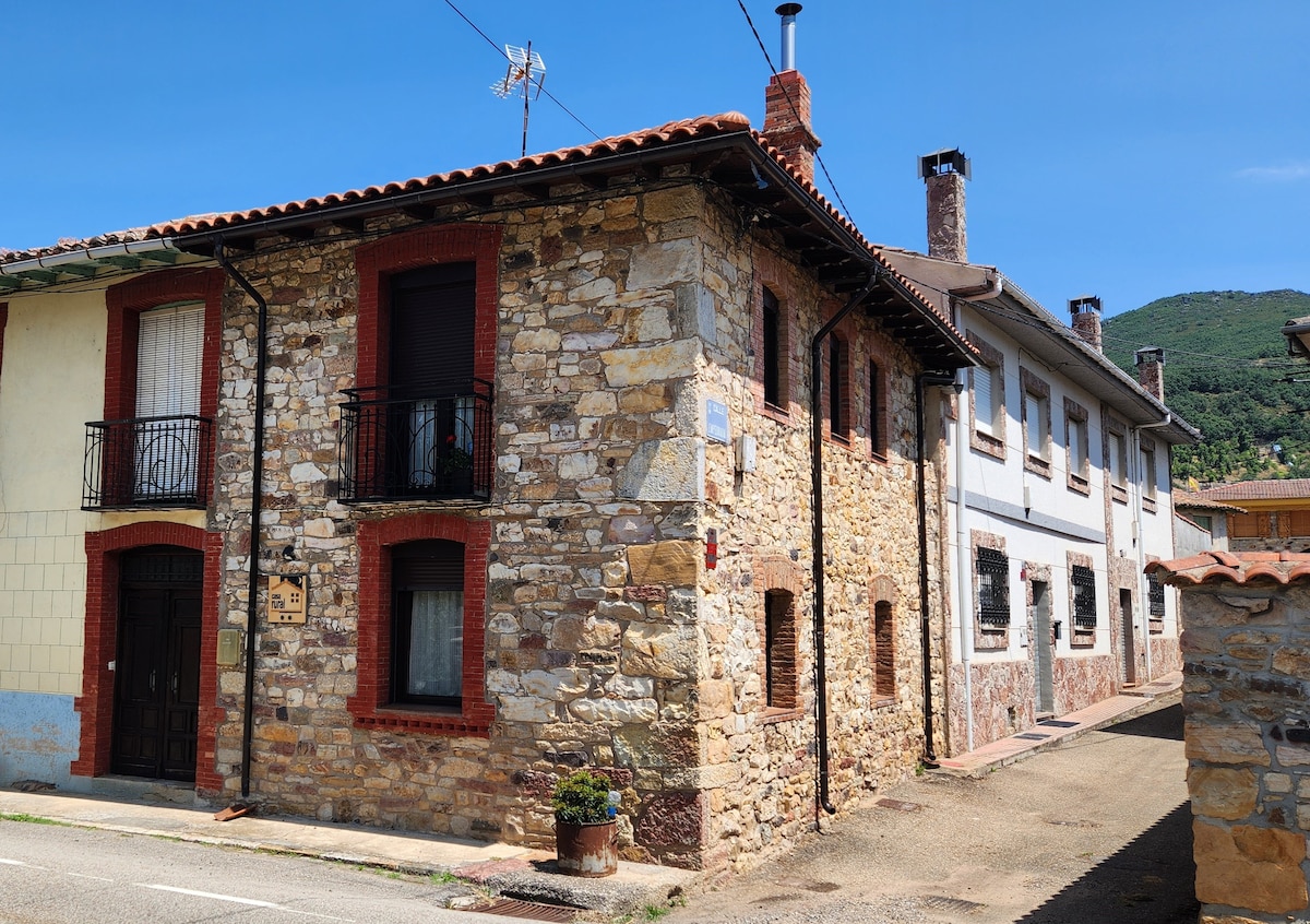 A charming stone house with a rustic exterior is depicted, showcasing a blend of light and dark stones. The building features multiple windows adorned with decorative balconies and a tiled roof. The setting reveals a peaceful street and surrounding green hills.