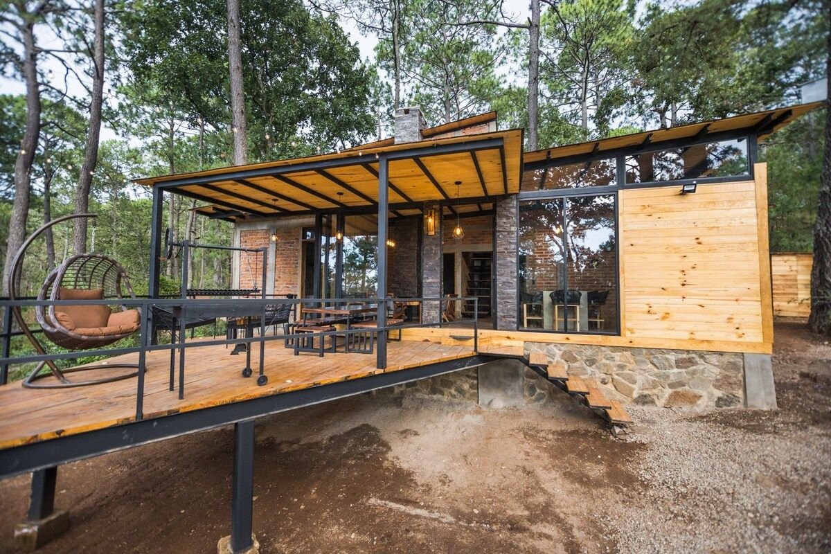 The exterior of the cabin is showcased with a wooden deck and a combination of stone and wood materials. Large glass windows provide a view into the living space. Outdoor seating arrangements are present, surrounded by lush trees, enhancing the connection with nature.