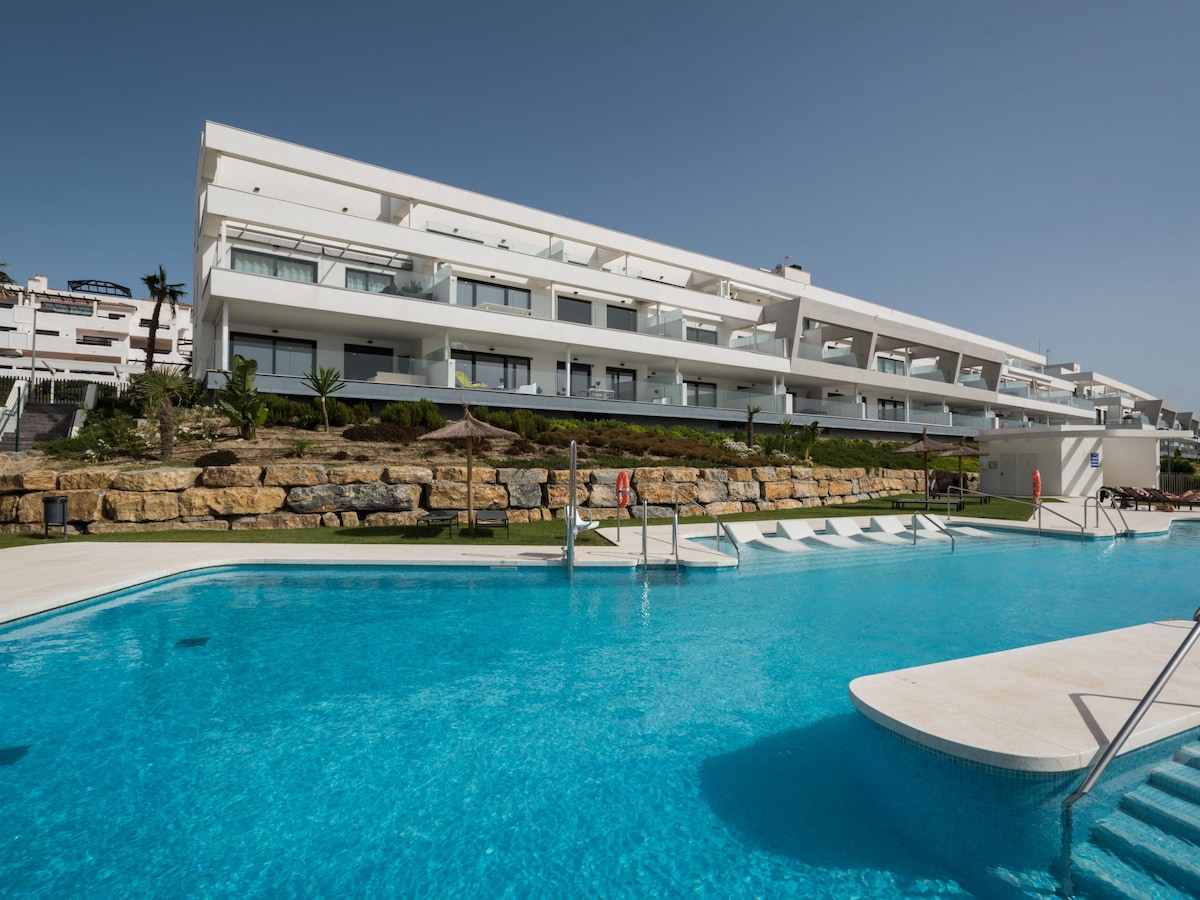 A modern apartment block is depicted with a clean white façade. A large angular swimming pool is shown in the foreground, surrounded by manicured lawns and landscaped rocks. Sun loungers and shaded areas are visible, contributing to the relaxing outdoor environment.