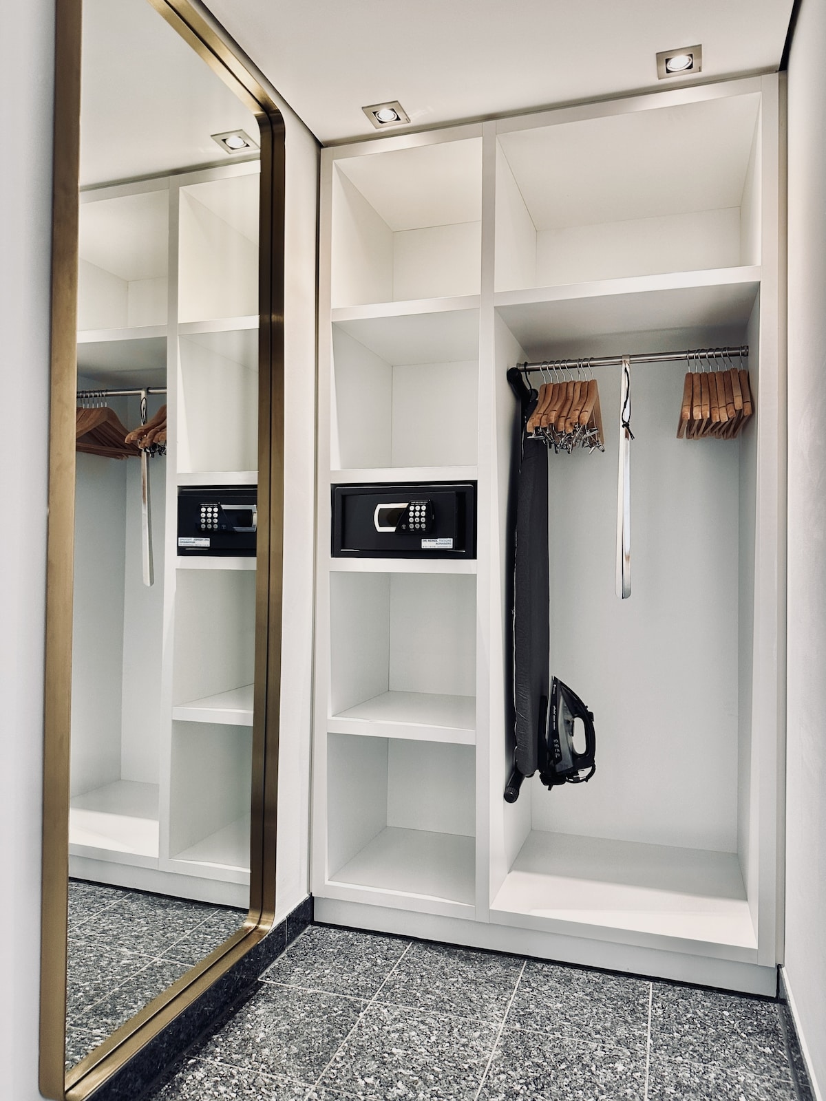 A spacious dressing area features a mirror alongside built-in shelves. A safe is prominently displayed, with hangers holding clothing items. An iron and ironing board are within easy reach. The flooring is adorned with dark tiles, contributing to a modern aesthetic.