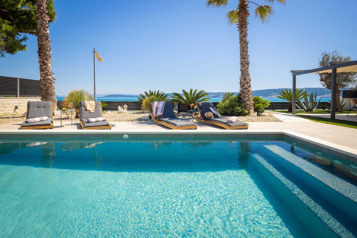 A serene pool area features a large heated swimming pool with sparkling turquoise water. Sun loungers are arranged around the pool, bordered by palm trees and lush greenery, offering views of the sea in the distance under a clear blue sky.