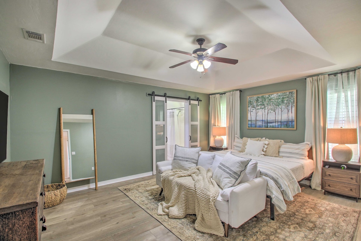 A spacious bedroom features a king bed with a light blanket and decorative pillows. A ceiling fan is installed above, and natural light enters through sheer curtains. A wall mirror and wooden dresser provide functional accents, while a soft area rug adds warmth to the space.
