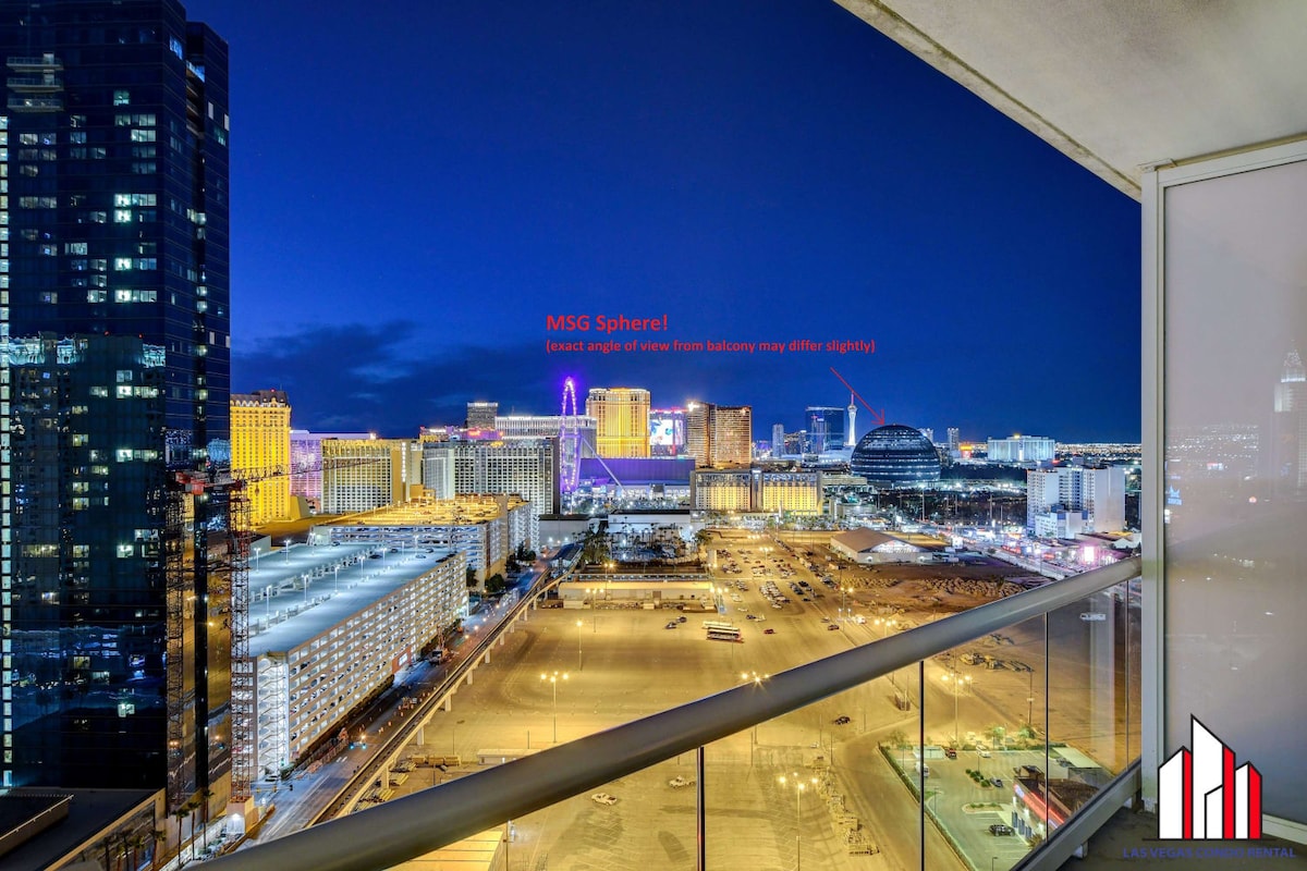 A cityscape view is captured from the balcony, showcasing the vibrant lights of the Las Vegas Strip and the MSG Sphere. The night sky serves as a backdrop for the illuminated buildings and attractions, providing a lively urban atmosphere.