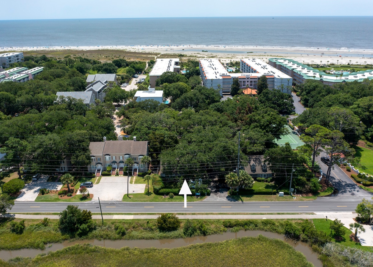 An aerial view captures a quaint condominium community surrounded by lush greenery, with the beach visible in the distance. The image highlights the proximity to the ocean and nearby developments, offering a sense of tranquility and easy access to beach activities.
