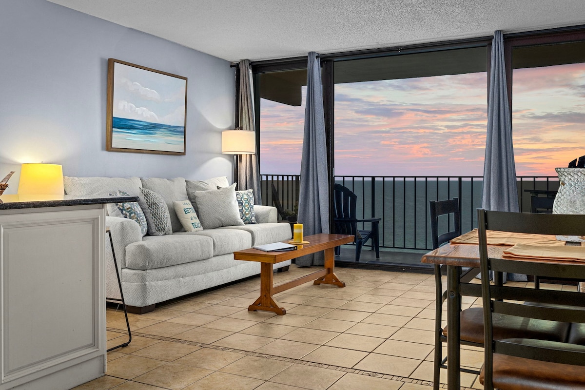 A light-filled living area features a comfortable, neutral-colored sofa with decorative pillows. A wooden coffee table sits in front, alongside a dining table with chairs. Sliding glass doors open to a balcony with views of the ocean and a vibrant sunrise, enhancing the serene atmosphere.