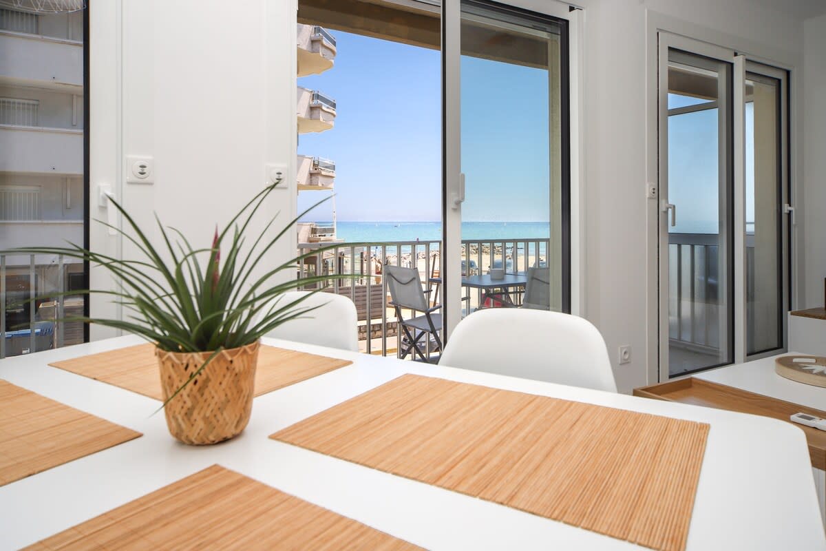 A bright dining area features a white table set with natural placemats. A potted plant adds a touch of greenery. Large glass doors open to a view of the sea, allowing natural light to flood the space.