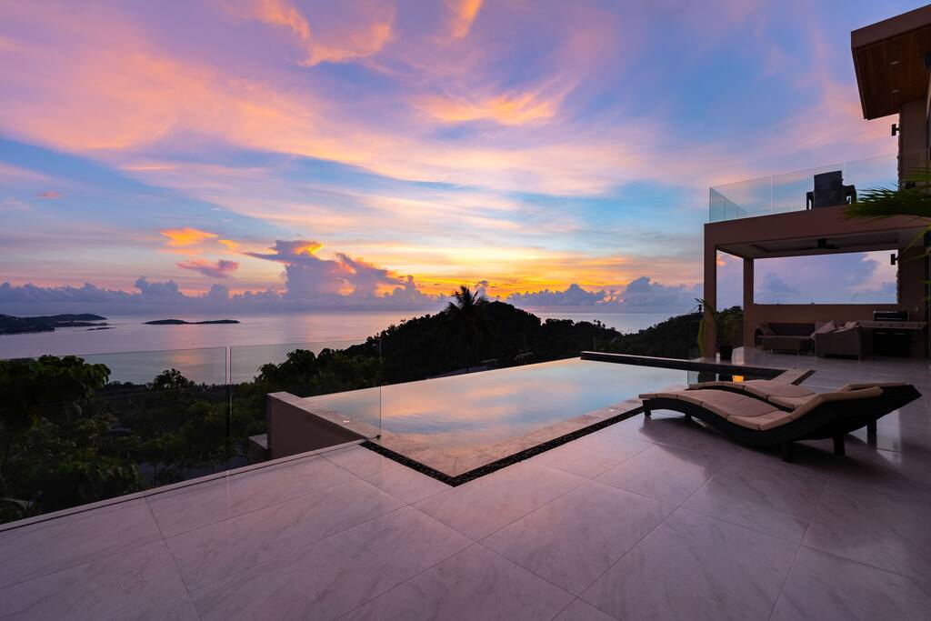 An infinity pool merges with the horizon, reflecting the vibrant colors of the sunset. The surrounding area features lush greenery and open space, inviting relaxation. Comfortable lounge chairs are positioned nearby, enhancing the tranquil ambiance of the outdoor living space.