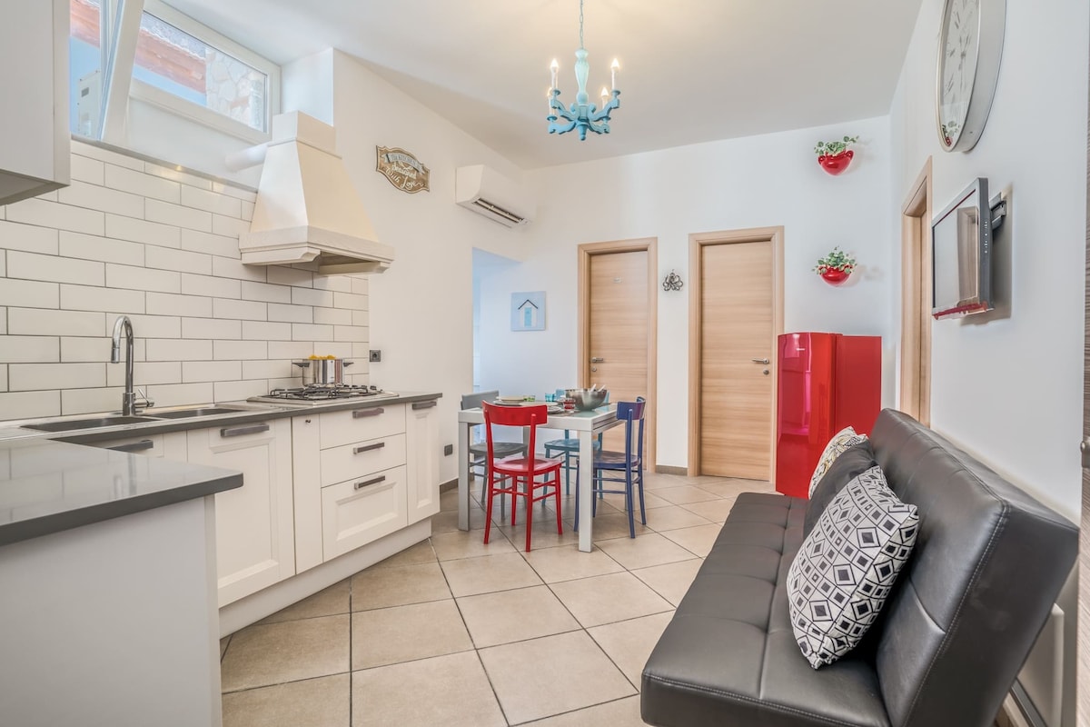 A modern kitchen space is highlighted with white cabinetry and a gray countertop. A red retro fridge stands against the wall, accompanied by a dining area featuring a small table surrounded by colorful chairs. A sofa is positioned nearby, adding comfort to the room.