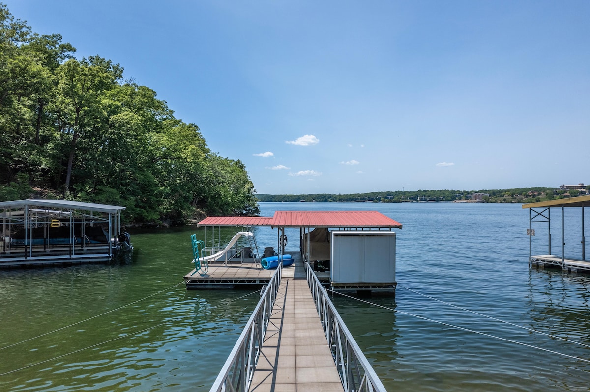 A wooden dock extends over calm waters leading to a covered boat slip. Lush green trees line the shore, providing shade. A blue kayak rests nearby, while the clear sky showcases scattered clouds, enhancing the serene lakeside atmosphere.