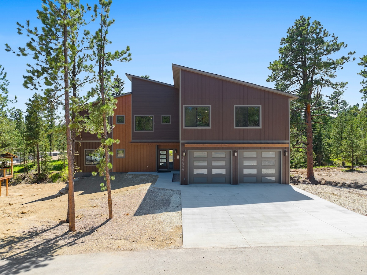 A modern two-story home with a combination of wood and metal siding is surrounded by tall pine trees. A wide driveway leads to a three-stall garage, and large windows provide ample natural light. The landscape features a mix of gravel and open space for additional outdoor activities.