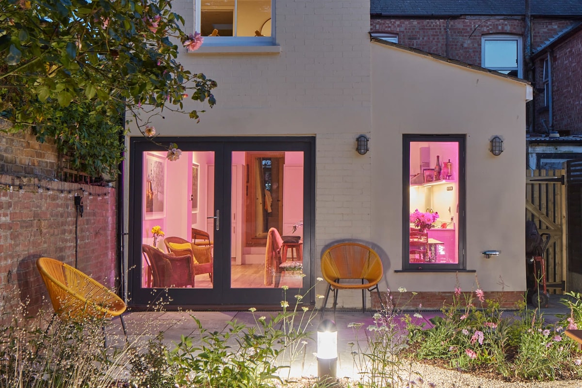 The exterior entrance is framed by colorful flower beds, featuring two stylish chairs positioned on a patio area. Large glass doors reveal a warm interior glow, creating a welcoming ambiance. The inviting space hints at a beautifully decorated living area visible through the doors.