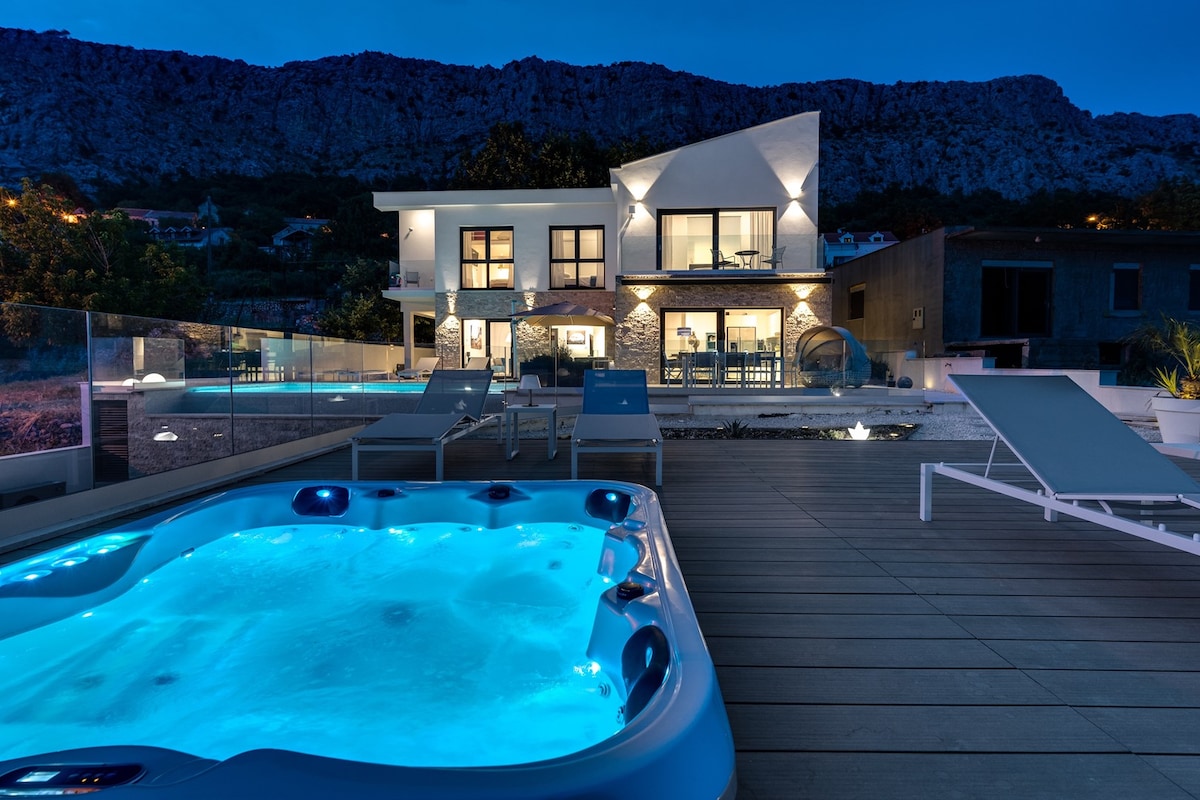 An inviting night view of the villa features an illuminated hot tub with blue water in the foreground. The modern structure showcases large windows and stone accents, reflecting soft lighting. Surrounding areas display sun loungers and a spacious deck, creating a serene atmosphere.
