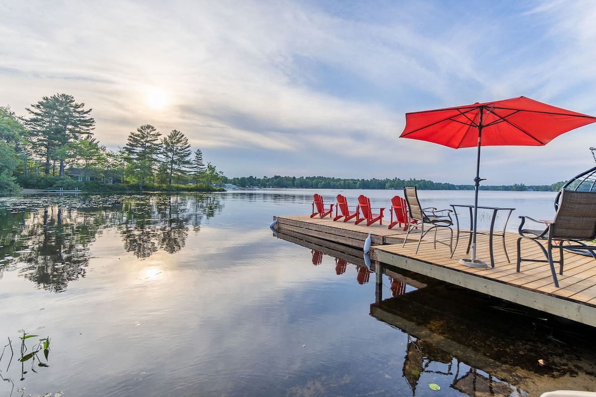 A spacious wooden dock extends over calm waters, featuring six red Adirondack chairs arranged under a large red umbrella. The tranquil scene is complemented by reflections of trees along the shoreline and a soft golden light from the setting sun.