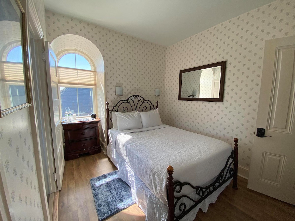 A bright room features a full bed framed in decorative ironwork. A large window allows natural light to fill the space, and a wooden dresser provides storage. A mirror is mounted on the wall, reflecting the room's soft floral wallpaper and warm wooden flooring.