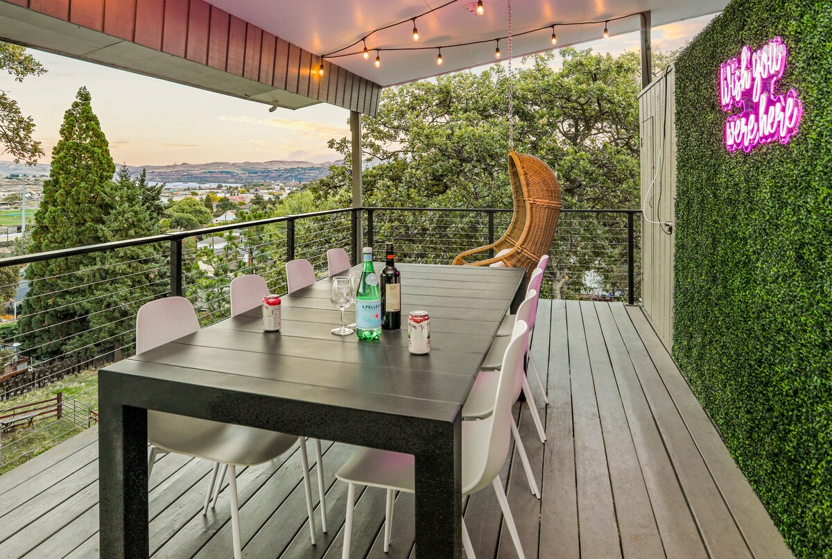 An outdoor deck offers a spacious dining table surrounded by white chairs, creating an inviting atmosphere. Soft lighting strung overhead enhances the space, while a neon sign adds a playful touch. A scenic view of the surrounding area can be appreciated from this elevated spot.