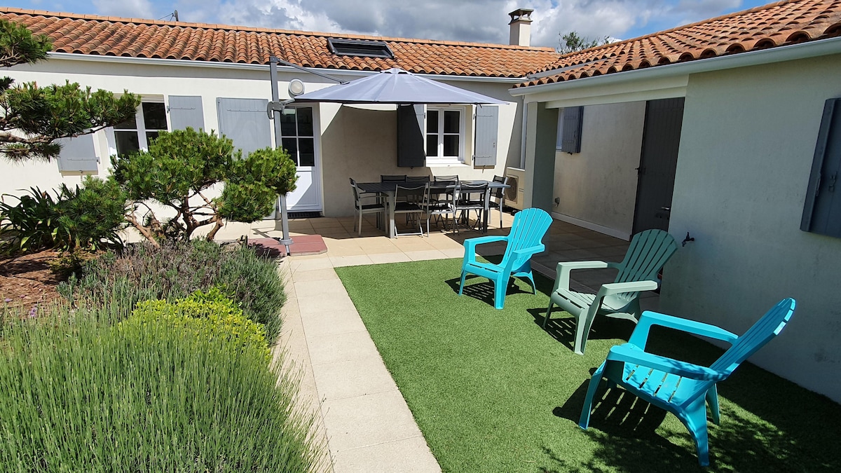 The outdoor area features a landscaped garden with artificial grass, showcasing two bright blue Adirondack chairs. A shaded dining space beneath a canopy is equipped with a table and chairs, surrounded by lavender plants and landscaped greenery.