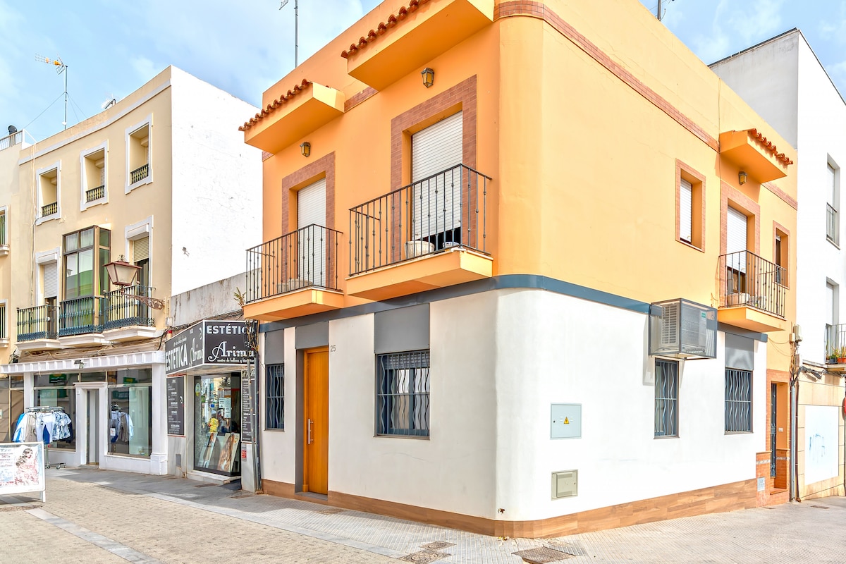 The exterior of a two-story building is displayed, featuring a vibrant orange facade and multiple balconies. The ground level includes a door and a series of windows with protective bars. A storefront is visible next door, complemented by a clear blue sky.