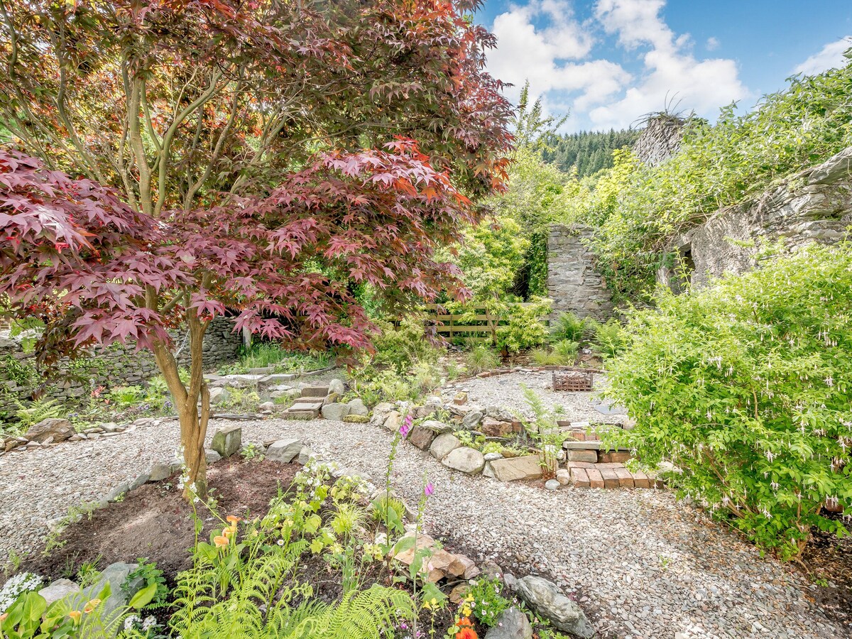 A tranquil garden area features a variety of lush greenery, including a vibrant red maple tree. Stone walls remain from a historic bakehouse, surrounded by winding paths and colorful flowers. The scene is enhanced by natural light filtering through trees and open spaces.