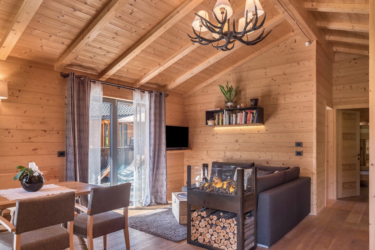 A cozy living room features wooden walls and a ceiling with exposed beams. A comfortable sofa is positioned near a modern fireplace, with decorative logs displayed below. A dining table with four chairs is also present, and large windows open to the outdoor terrace.