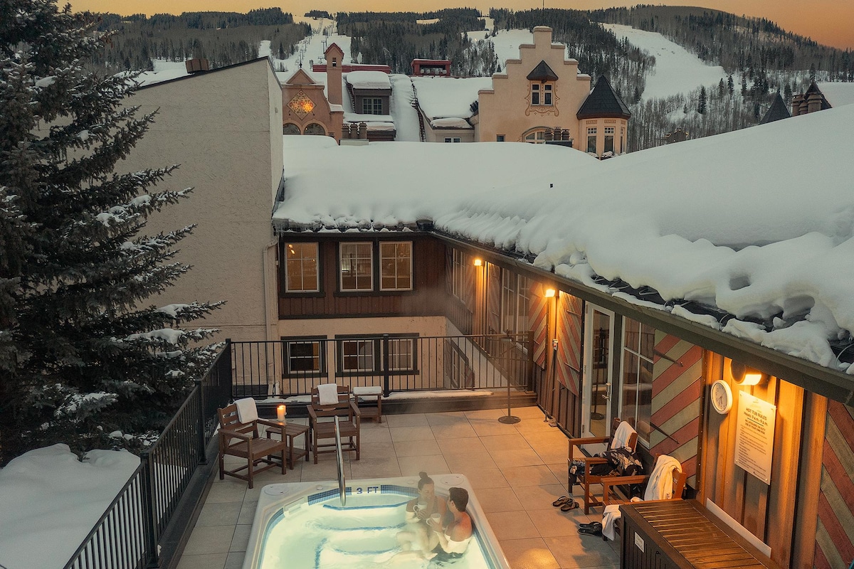 A rooftop hot tub is surrounded by snow-covered rooftops, providing a serene view of the Vail landscape during twilight. Warm lighting illuminates the space, creating a cozy ambiance. Several lounge chairs are positioned nearby for relaxation.