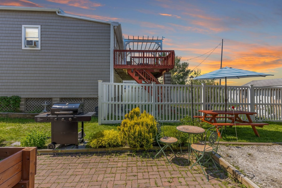 An outdoor patio area is featured, with a gas grill positioned next to a small dining table and chairs. A picnic table and large umbrella are visible in the background, surrounded by green grass and a white fence. The sky displays hues of orange and pink at sunset.