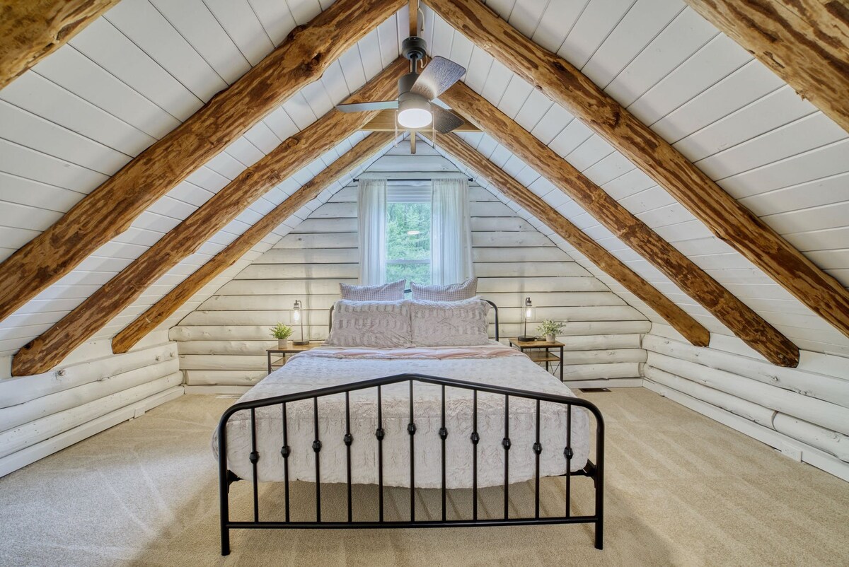 An inviting bedroom is depicted, featuring a spacious bed with a light-colored blanket. Natural light filters through a window, framed by sheer curtains. Exposed wooden beams create a rustic atmosphere, enhancing the room's character. Two small plants are positioned on nightstands beside the bed.