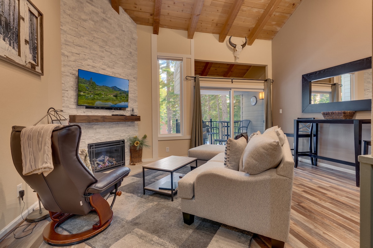 A cozy living room features a modern gas fireplace below a flat screen TV. Natural light filters in from large windows and a sliding glass door, which leads to a covered deck. The space includes a comfortable sofa, a plush armchair, and a coffee table.