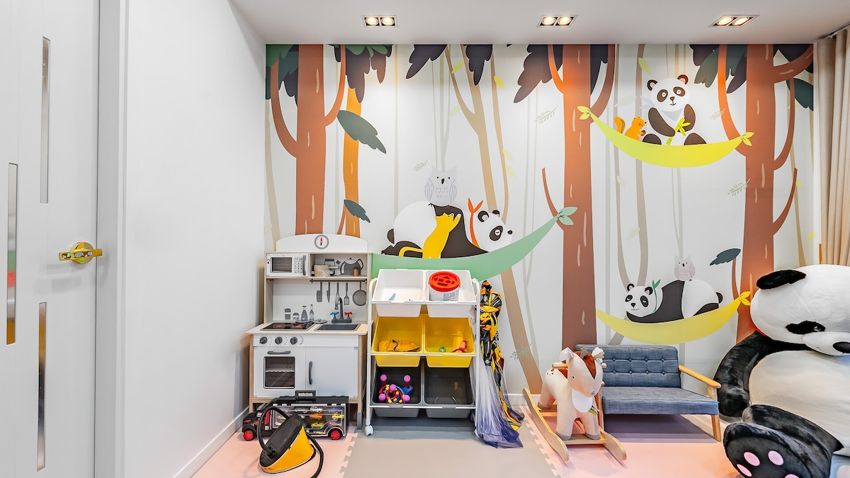 A playful children's room features a vibrant mural of trees with panda and other animal illustrations. A cozy seating area includes a blue sofa, while a toy kitchen and storage bins are organized for easy access. Soft toys provide additional comfort and fun.