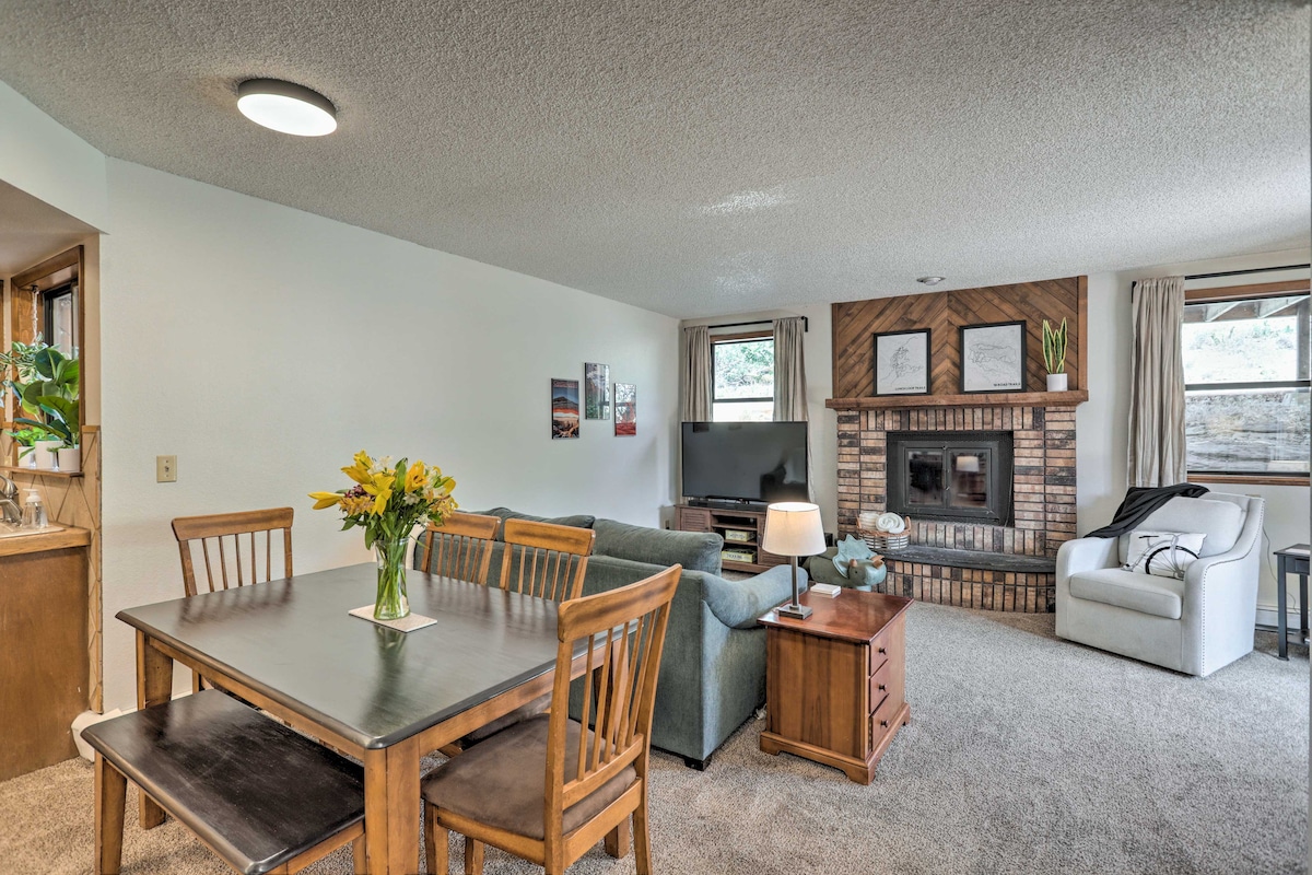 An open living area displays a dining table with wooden chairs and a vase of flowers at its center. A comfortable seating arrangement faces a smart TV, with a decorative brick fireplace adding warmth. Natural light filters through the curtains of the adjacent windows.