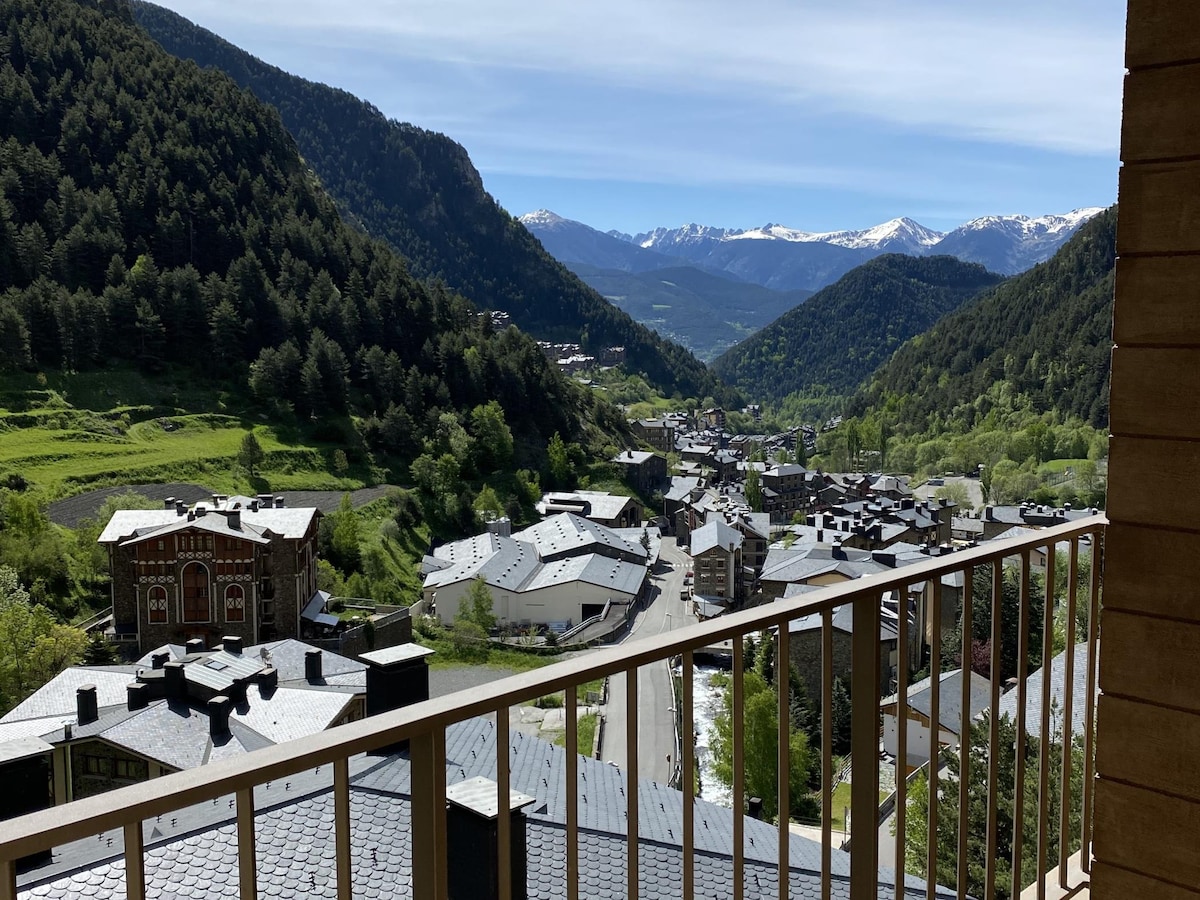 A panoramic view from the balcony reveals expansive green valleys and tall mountains, highlighted by patches of snow. The village of Arinsal is nestled below, featuring a mix of rooftops and roads that curve through the landscape.