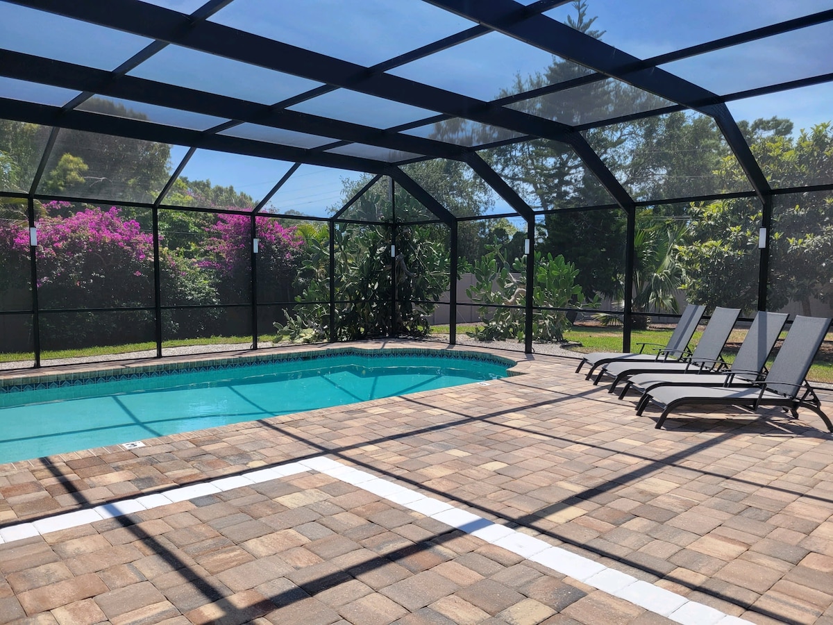 An enclosed patio features a tranquil swimming pool surrounded by patterned stone flooring. Sun loungers are positioned nearby, and lush greenery is visible beyond the screen, creating a serene outdoor space for relaxation and enjoyment.