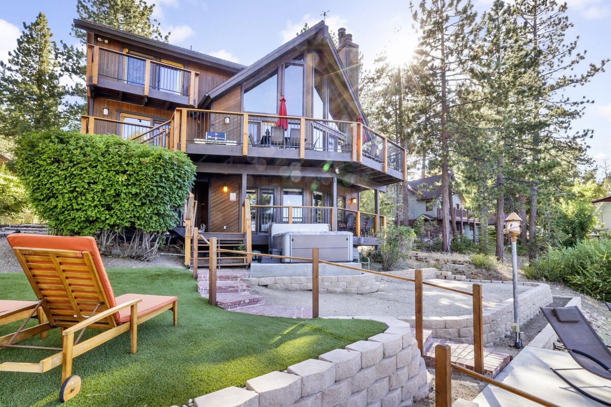 A spacious lakefront chalet is showcased, featuring a large deck with a hot tub and lounge chairs. Lush greenery surrounds the property, with steps leading down to a lakeside area. Sunlight bathes the home, highlighting its outdoor spaces and inviting atmosphere.