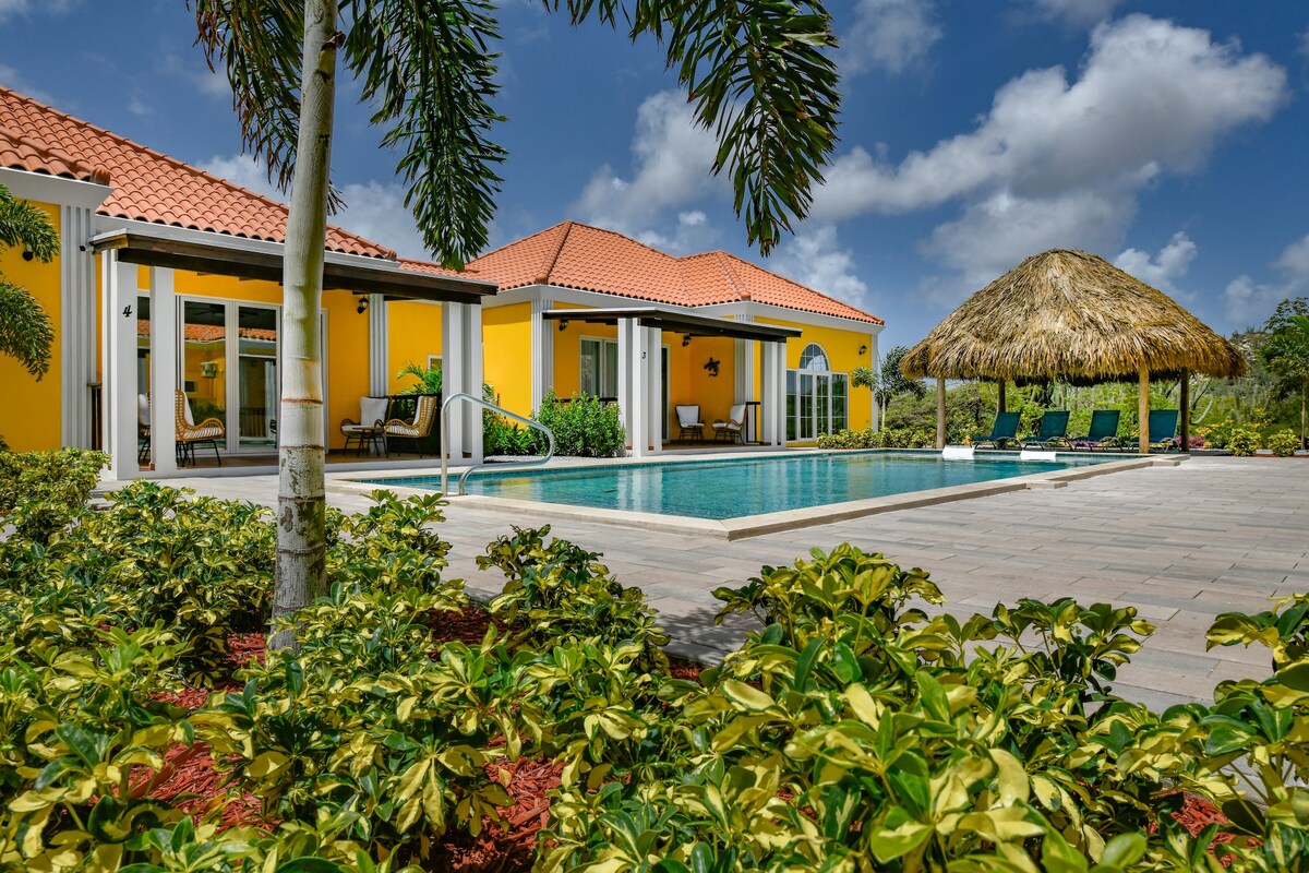 A vibrant yellow villa is set against a clear blue sky, featuring a serene pool and a thatched-roof gazebo. Lush green landscaping surrounds the outdoor area, creating a tropical atmosphere. Large glass doors offer views of the inviting space, emphasizing the indoor/outdoor connection.