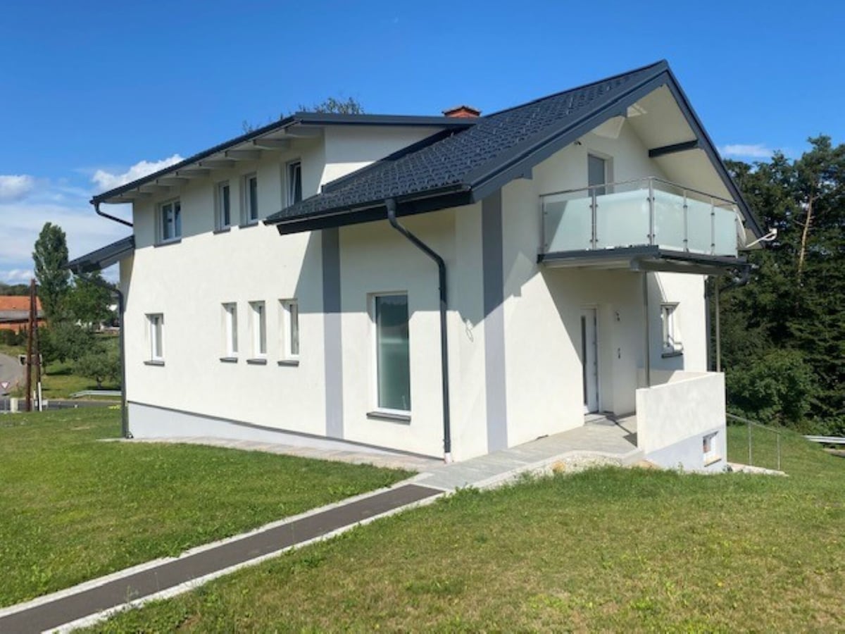The exterior of the house showcases a modern design with a white façade and a dark roof. A balcony is visible on the first floor, and large windows offer natural light. The surrounding lawn is well-maintained, contributing to the tranquil setting.