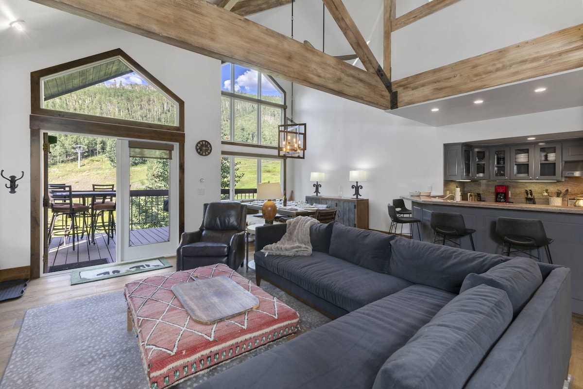 An open living area features a sectional couch, accented with a patterned area rug. Large windows allow natural light to fill the space, revealing views of the surrounding landscape. A cozy dining area and a modern kitchen with bar seating are also visible in the background.