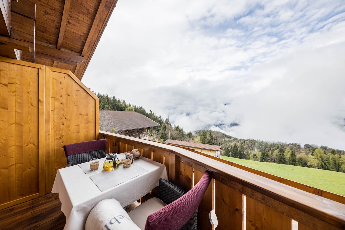 A private balcony is depicted, featuring a wooden railing and a round table set with a tablecloth. Two chairs are placed on either side of the table. The background showcases a view of rolling hills and clouds, creating a serene atmosphere.