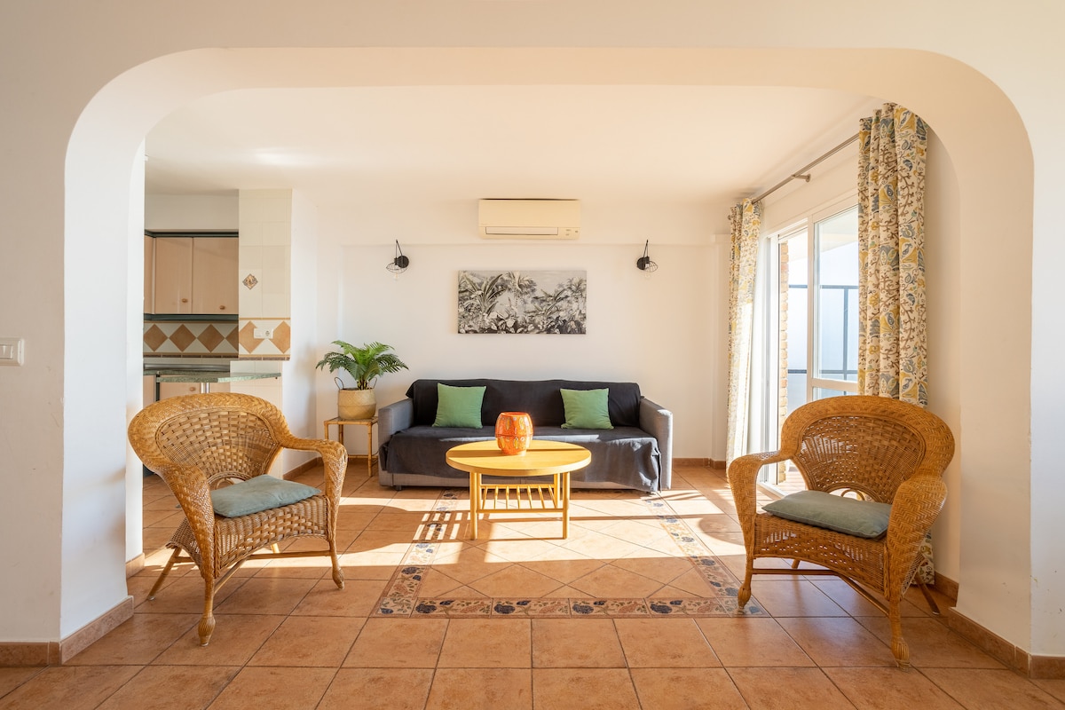 A bright living room features a sofa bed accompanied by two wicker chairs, arranged around a round coffee table. Natural light floods in through large windows with soft curtains, creating a welcoming atmosphere. A decorative wall art piece complements the neutral color scheme.