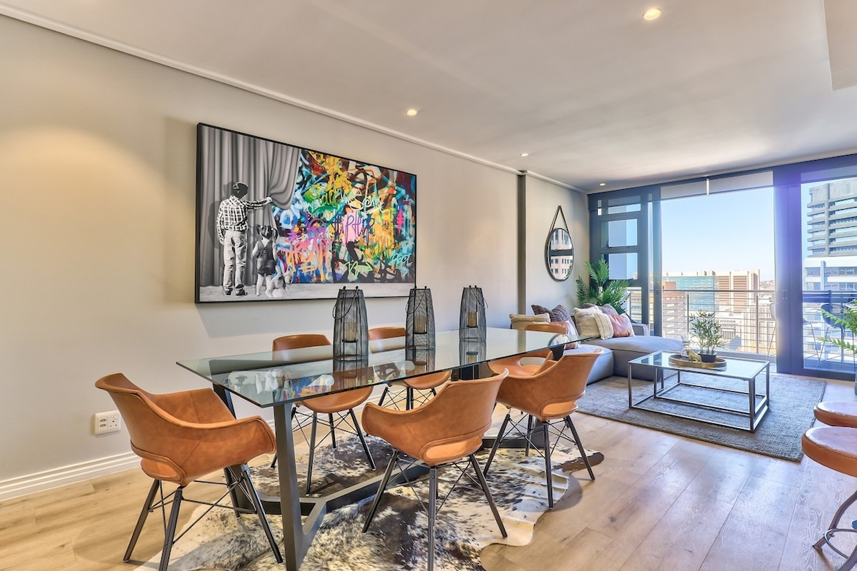 A dining area features a sleek glass table surrounded by tan upholstered chairs. A vibrant piece of contemporary art adorns the wall, while natural light enters through large windows, illuminating the space. A light-toned wooden floor adds warmth to the modern decor.