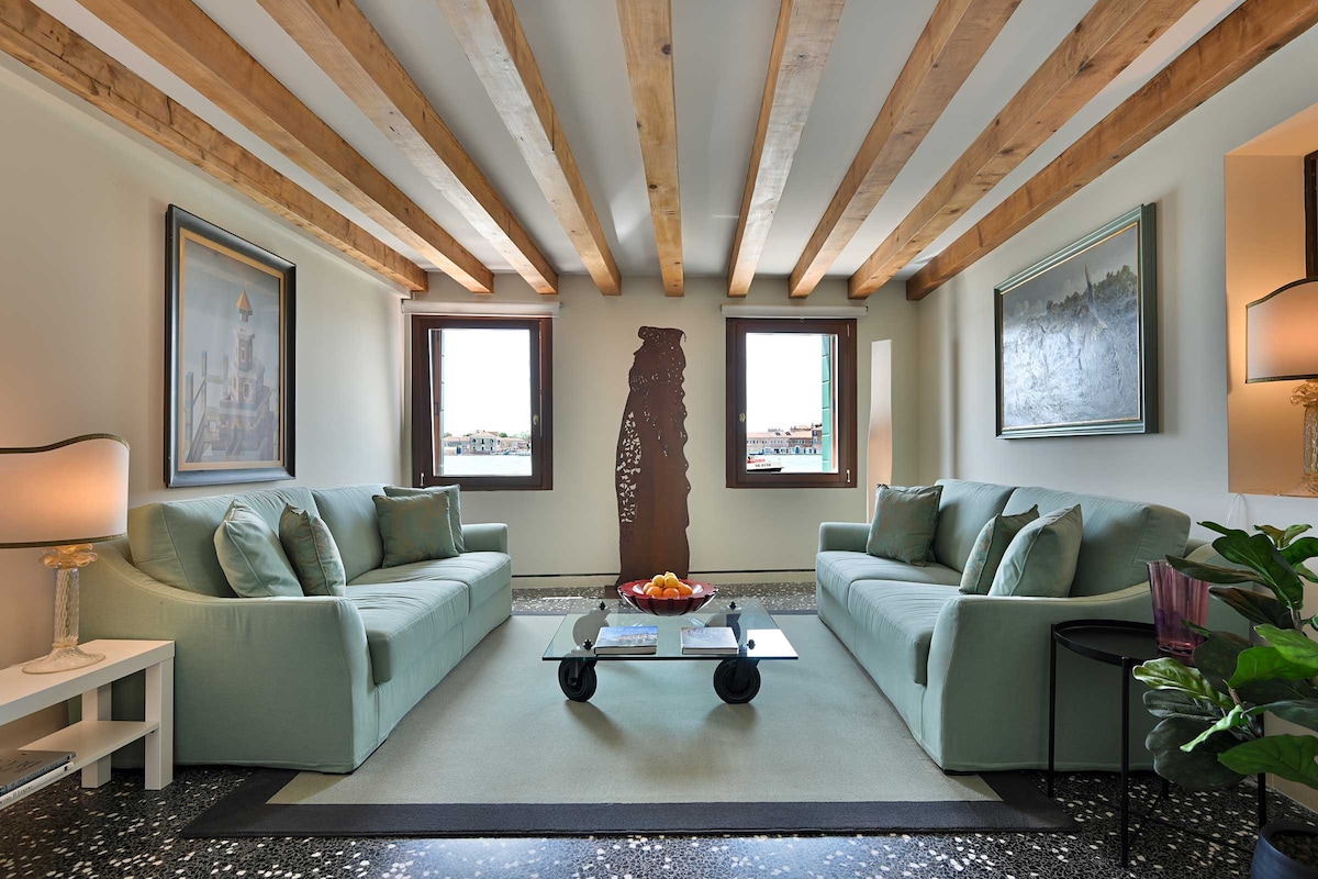 A bright living area features two light green sofas positioned around a coffee table with a dark base. Natural light enters through two windows, enhanced by exposed wooden beams above. Decorative art pieces adorn the walls, complemented by a potted plant in one corner.