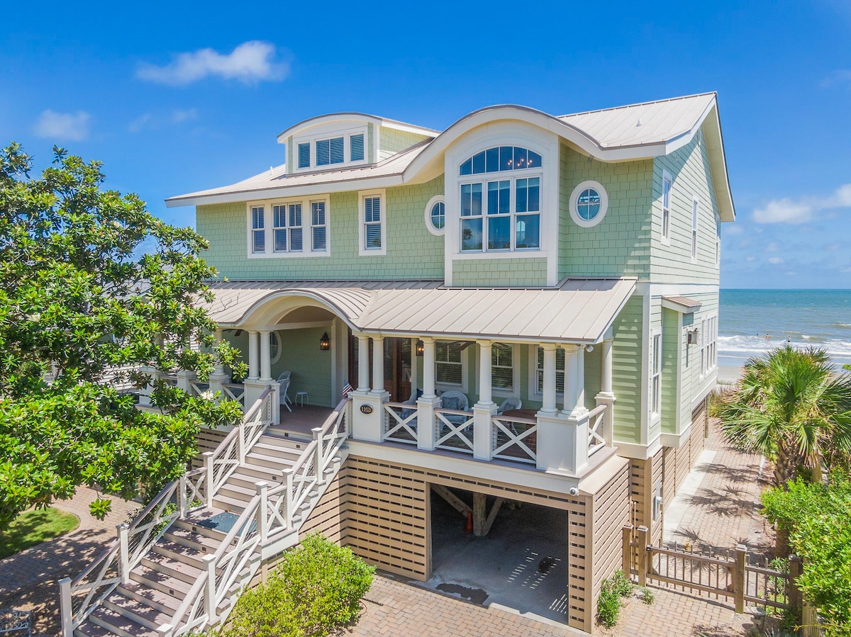 An oceanfront house with a charming design features large windows and a covered front porch. A pathway leads up to the entrance, framed by lush greenery. Underneath, parking space is visible, allowing easy access to the beach just beyond.