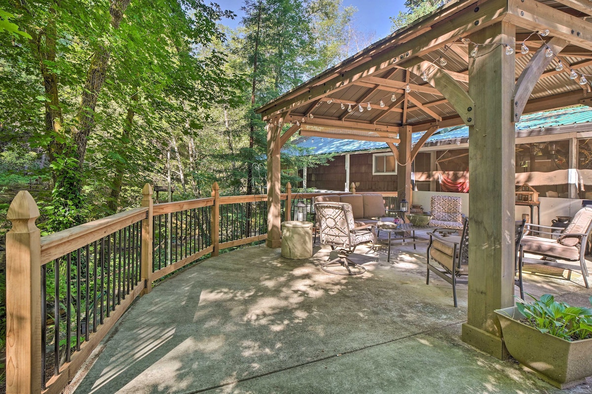 An outdoor living area is shaded by a gazebo, featuring multiple lounge chairs arranged around a stone table. Wood and metal railings enhance the space, which overlooks a lush backdrop of trees. Soft lighting is visible above, creating a welcoming ambiance.
