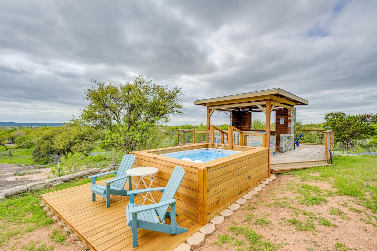 An inviting plunge pool is surrounded by a wooden deck, complemented by two light blue Adirondack chairs and a small table. A covered cabana is located nearby, offering shaded seating and a panoramic view of the lush greenery and distant hills.