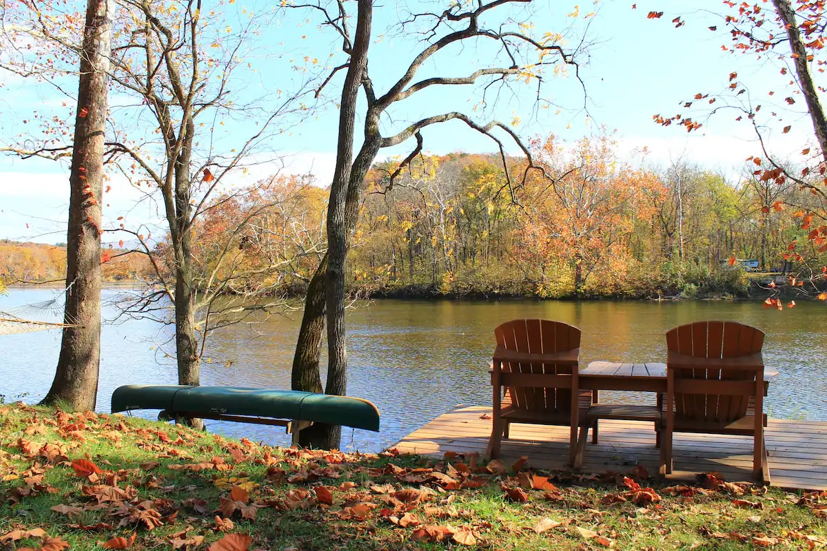 A wooden deck overlooks the Shenandoah River, featuring two Adirondack chairs accompanied by a small table. A green canoe is secured nearby, and vibrant autumn foliage enhances the scenic riverside setting, with trees casting dappled shadows on the grass.