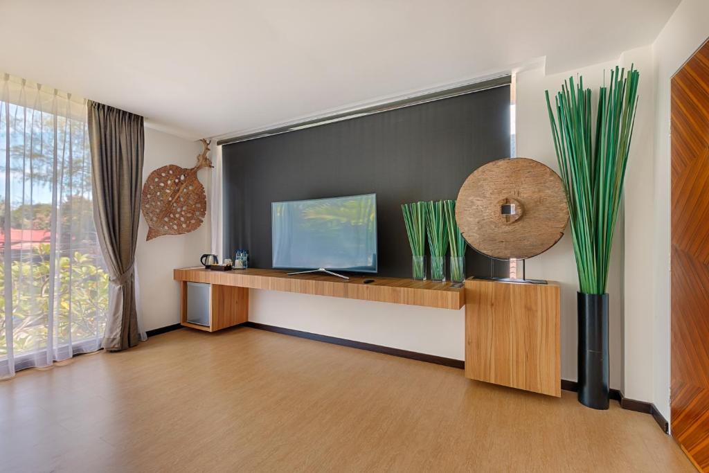 A stylish living area features a wall-mounted television above a wooden console. Decorative elements include large round appliques and tall green plants. Natural light filters through sheer curtains, while a wooden accent wall adds texture to the space.