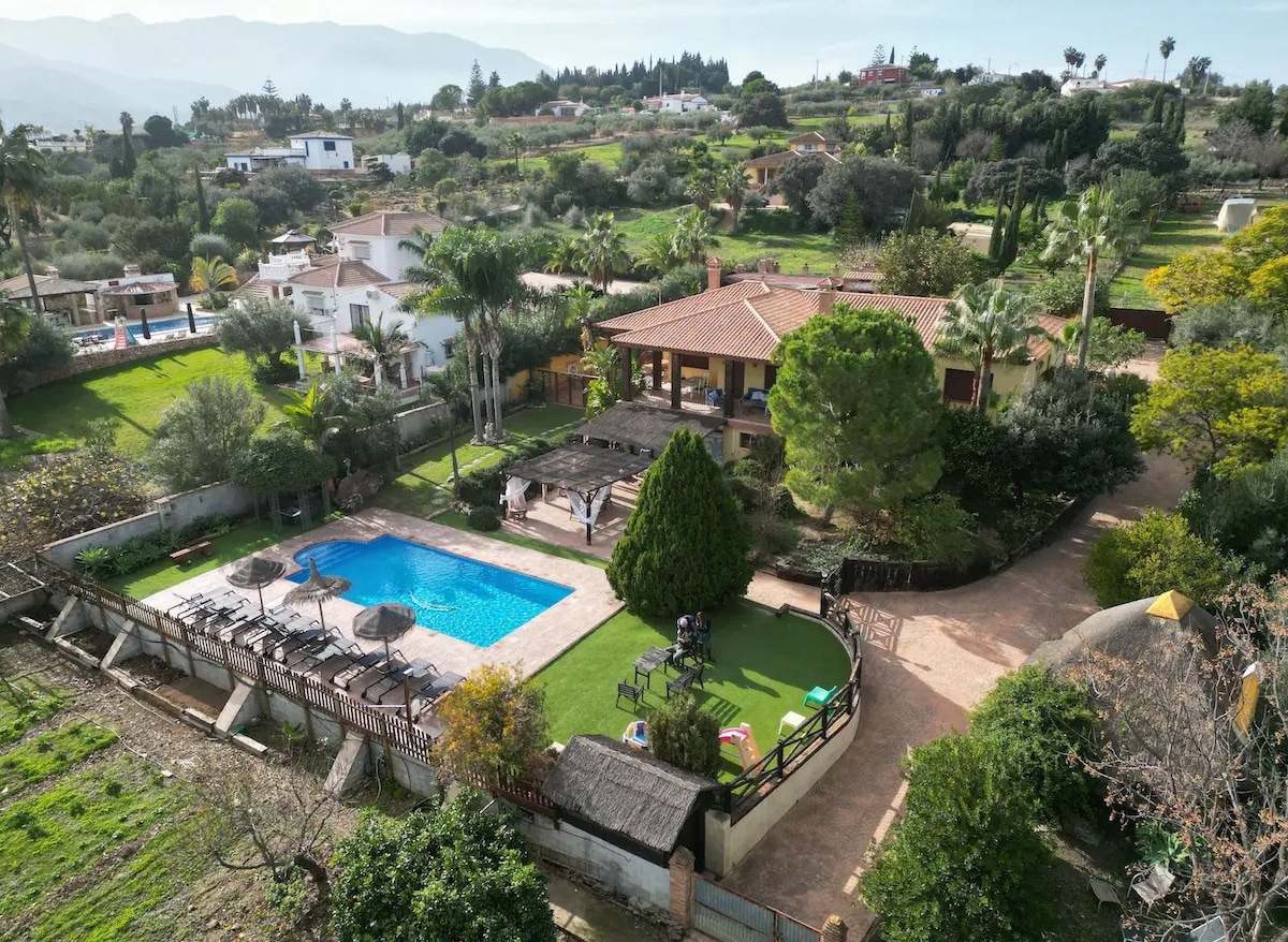 An aerial view of the villa reveals a spacious outdoor area surrounded by lush greenery. A swimming pool is positioned centrally, complemented by sun loungers and shaded areas. The building showcases a terracotta roof and is set against a backdrop of rolling hills, enhancing the serene environment.