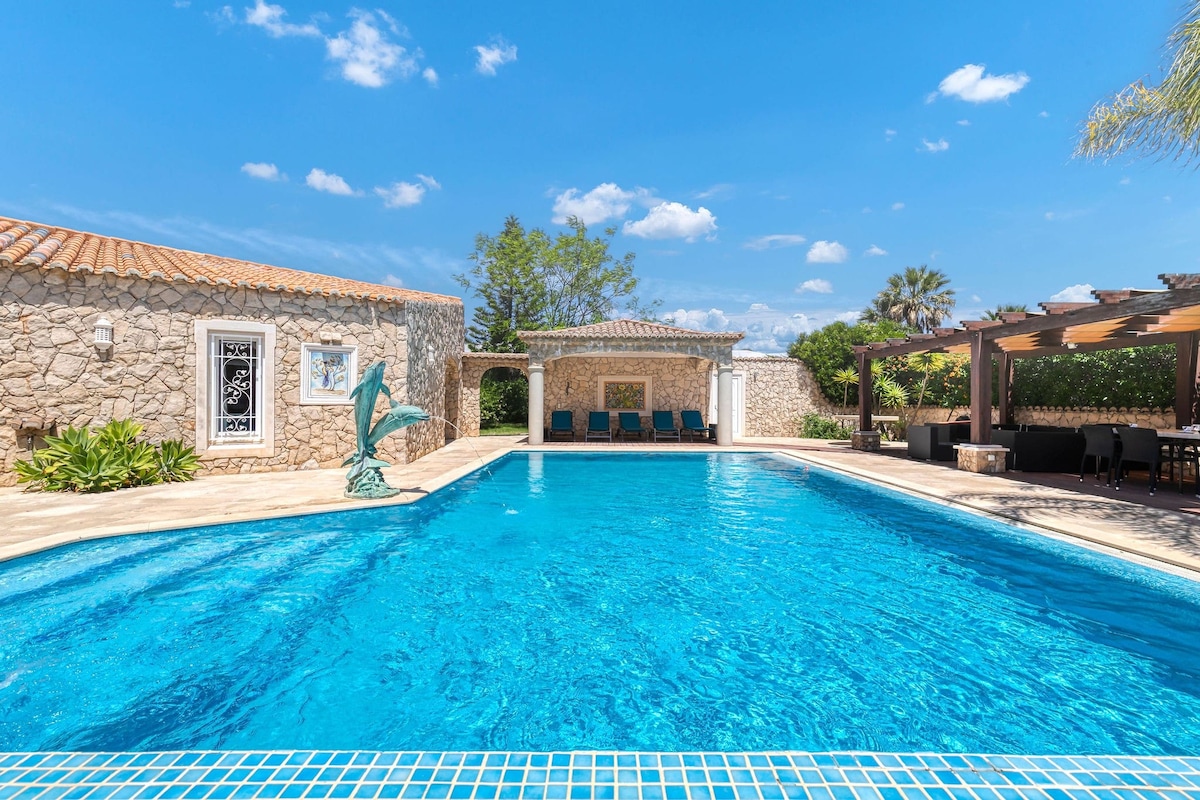 A large swimming pool is set against a backdrop of natural stone walls and vibrant greenery. Comfortable loungers are arranged along the poolside, with shaded areas provided by a nearby pergola. The bright blue sky enhances the inviting atmosphere of the outdoor space.