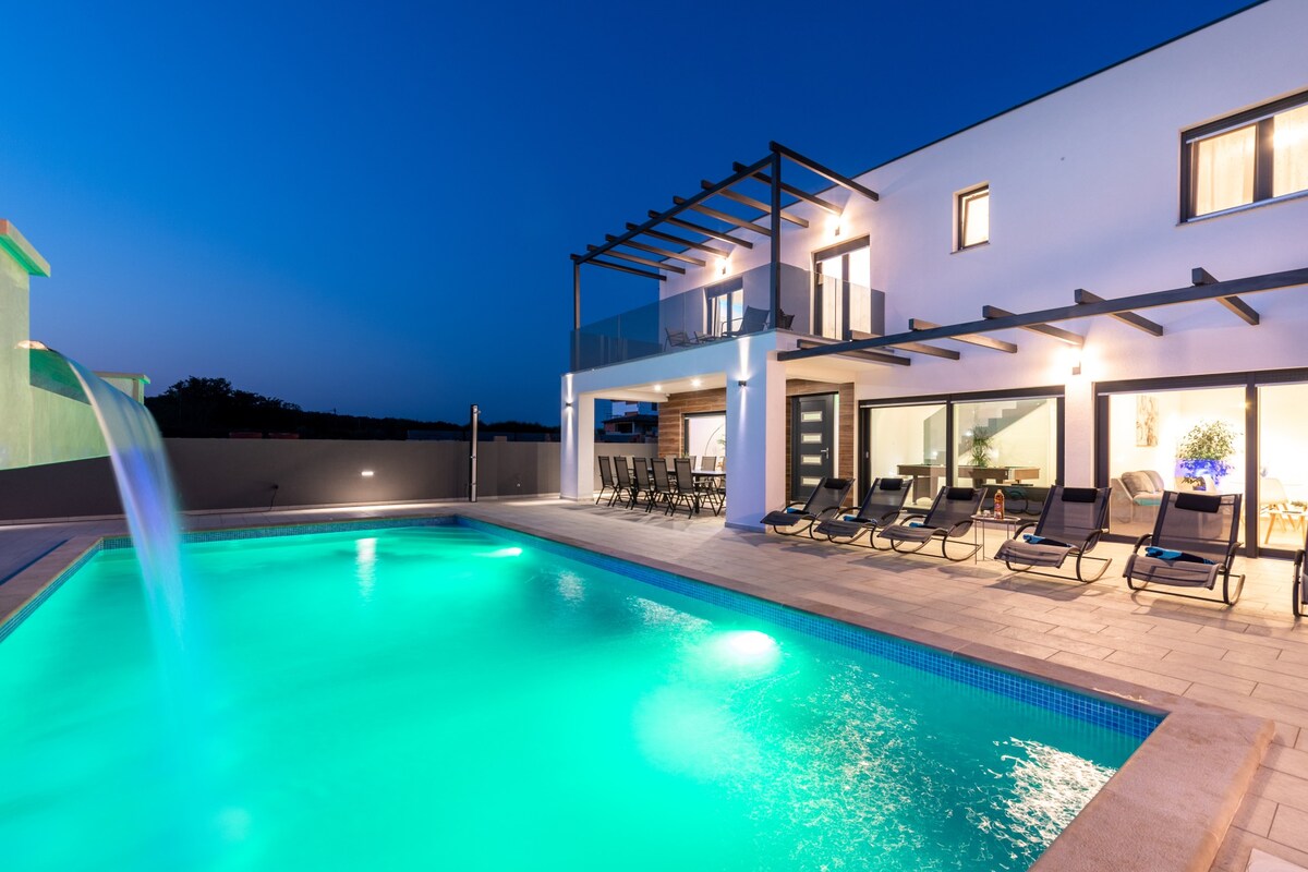 A modern villa exterior is illuminated under the evening sky, showcasing a large heated swimming pool with a waterfall feature. Comfortable lounge chairs are arranged around the pool, and expansive glass doors provide views into the interior spaces.