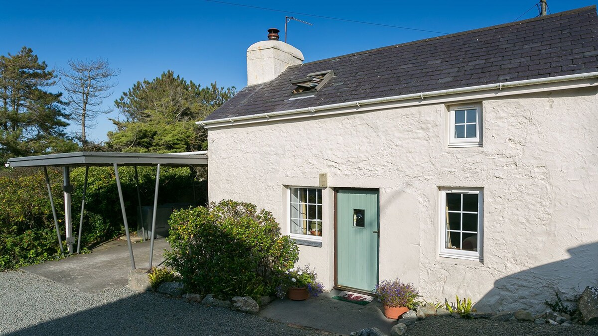The exterior of a quaint holiday cottage displays a light-coloured stone façade and a welcoming green door. A covered entrance area provides shelter, while flower pots filled with greenery add charm. Surrounding trees and shrubs enhance the cottage's natural setting.